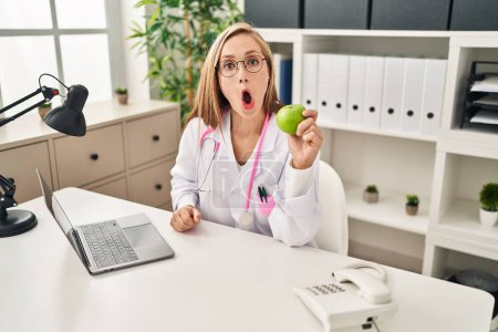 Téléchargez les photos : Jeune femme blonde médecin tenant pomme verte à la clinique effrayée et étonnée avec la bouche ouverte pour surprise, visage incrédule - en image libre de droit