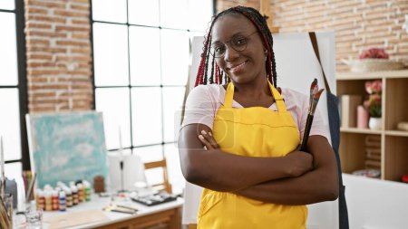 Foto de Confiada artista afroamericana con una sonrisa brillante, de pie en su estudio de arte, pinceles en la mano, brazos cruzados - Imagen libre de derechos