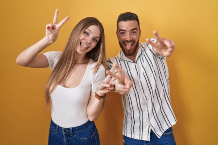 Téléchargez les photos : Jeune couple debout sur fond jaune souriant avec la langue montrant les doigts des deux mains faisant signe de victoire. numéro deux. - en image libre de droit