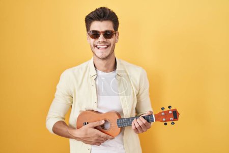 Foto de Joven hombre hispano jugando ukelele sobre fondo amarillo sonriendo y riendo en voz alta porque divertido chiste loco. - Imagen libre de derechos