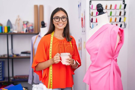Téléchargez les photos : Jeune belle femme hispanique tailleur boire du café debout par mannequin au magasin de tailleur - en image libre de droit