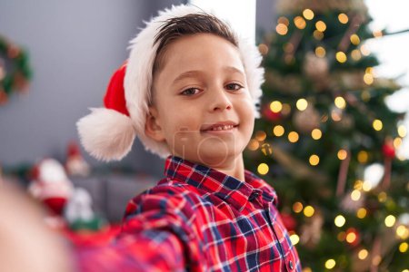 Téléchargez les photos : Adorable garçon hispanique faire selfie par caméra debout près de l'arbre de Noël à la maison - en image libre de droit