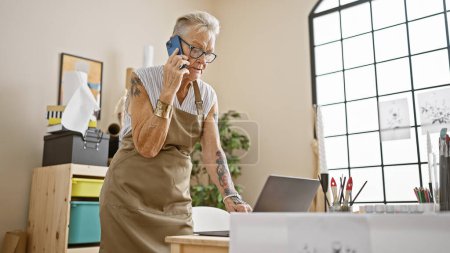 Photo for Confident, grey-haired senior woman artist adeptly multitasks, speaking on smartphone while using laptop in her inspiring art studio interior. - Royalty Free Image