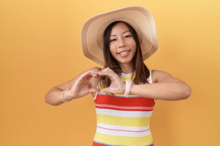 Foto de Mujer china de mediana edad con sombrero de verano sobre fondo amarillo sonriendo en el amor haciendo forma de símbolo del corazón con las manos. concepto romántico. - Imagen libre de derechos