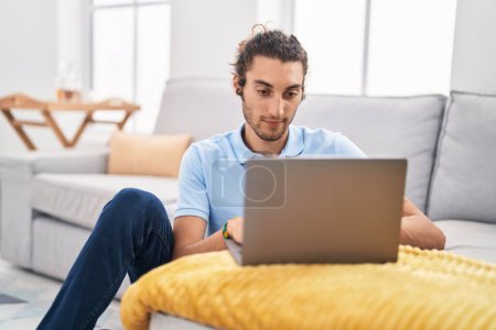 Téléchargez les photos : Jeune homme hispanique utilisant un ordinateur portable assis sur le sol à la maison - en image libre de droit