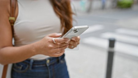 Foto de Mujer hispana joven usando sus manos para enviar un mensaje de texto en su teléfono al aire libre en el corazón de la calle de la ciudad, conectándose al mundo digital. - Imagen libre de derechos