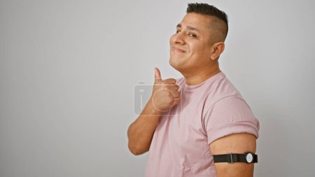 Foto de Joven latino confiado sonriendo con el pulgar hacia arriba, relajándose mientras monitorea la diabetes con el sensor de brazo - de pie aislado sobre fondo blanco - Imagen libre de derechos