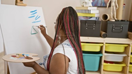 Téléchargez les photos : Belle artiste afro-américaine ornant des tresses dessinant passionnément dans un studio d'art - en image libre de droit