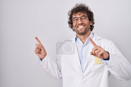 Foto de Joven hispano vistiendo uniforme médico sonriendo y mirando a la cámara apuntando con dos manos y dedos hacia un lado. - Imagen libre de derechos