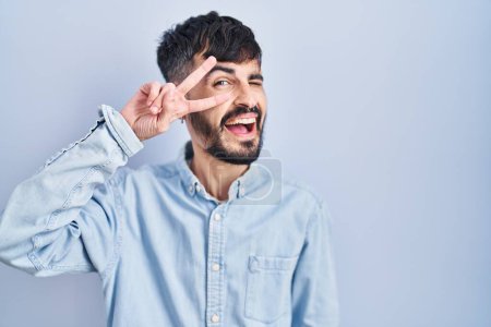 Téléchargez les photos : Jeune homme hispanique avec la barbe debout sur fond bleu faisant symbole de paix avec les doigts sur le visage, souriant joyeux montrant la victoire - en image libre de droit