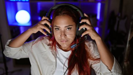 Photo for Focused young redhead woman streamer, engrossed in intense gaming session, in a serious virtual face-off, headset on, encapsulated in the dimly-lit indoors of her home gaming room - Royalty Free Image
