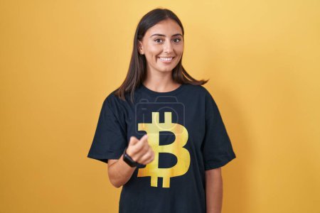 Téléchargez les photos : Jeune femme hispanique portant Bitcoin t-shirt signe venir ici geste avec la main invitant accueillant heureux et souriant - en image libre de droit