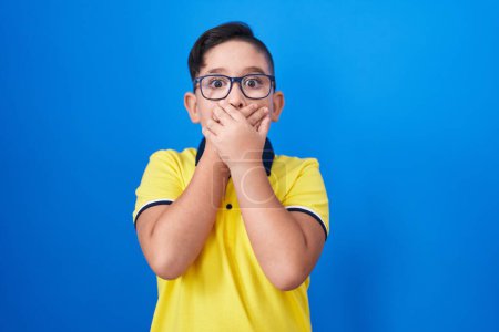 Foto de Young hispanic kid standing over blue background shocked covering mouth with hands for mistake. secret concept. - Imagen libre de derechos
