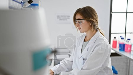 Foto de Absorto en la investigación médica, joven, hermosa mujer rubia científica, enfocada y seria, trabajando en la computadora en el laboratorio - Imagen libre de derechos