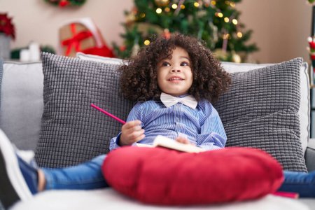 Foto de Niño afroamericano dibujando en un cuaderno sentado en un sofá junto al árbol de Navidad en casa - Imagen libre de derechos