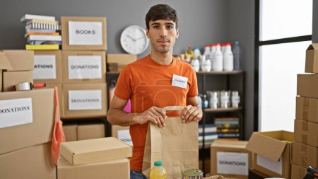 Foto de Joven hombre hispano guapo centrado en el voluntariado, sosteniendo bolsa de papel en un centro de caridad en el interior - Imagen libre de derechos