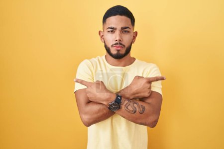 Foto de Young hispanic man standing over yellow background pointing to both sides with fingers, different direction disagree - Imagen libre de derechos