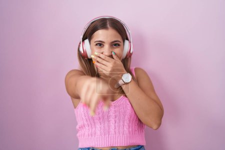 Téléchargez les photos : Jeune femme blonde écoutant de la musique à l'aide d'écouteurs se moquant de vous, pointant du doigt la caméra avec la main sur la bouche, expression de honte - en image libre de droit