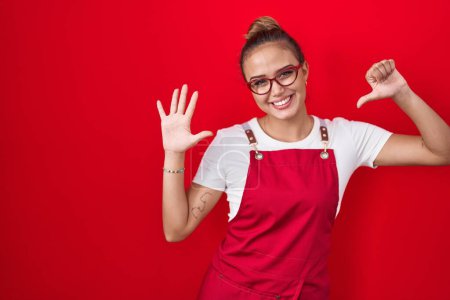 Téléchargez les photos : Jeune femme hispanique portant tablier de serveuse sur fond rouge montrant et pointant vers le haut avec les doigts numéro six tout en souriant confiant et heureux. - en image libre de droit