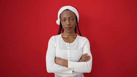 Foto de Mujer afroamericana con sombrero de navidad de pie con expresión seria y brazos cruzados gesto sobre fondo rojo aislado - Imagen libre de derechos