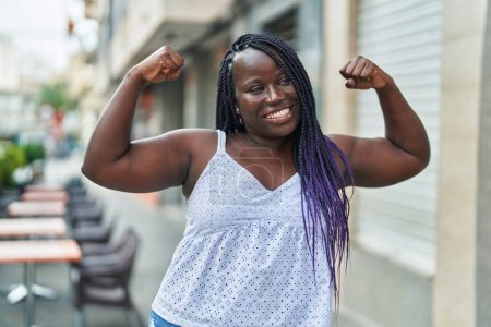 Foto de Mujer afroamericana sonriendo confiada haciendo un gesto fuerte con los brazos en la terraza de la cafetería - Imagen libre de derechos
