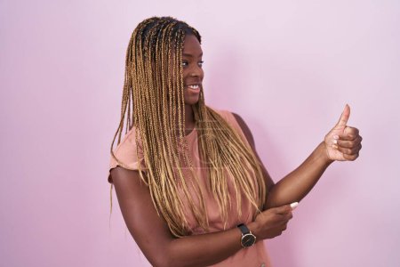 Téléchargez les photos : Femme afro-américaine avec les cheveux tressés debout sur fond rose regardant fier, souriant faire les pouces vers le haut geste sur le côté - en image libre de droit