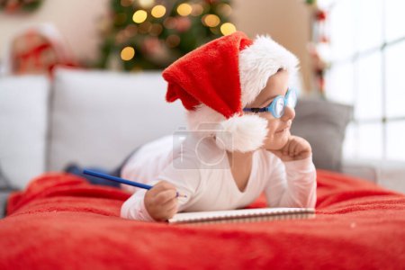 Photo for Adorable hispanic toddler lying on sofa by christmas tree writing on notebook at home - Royalty Free Image