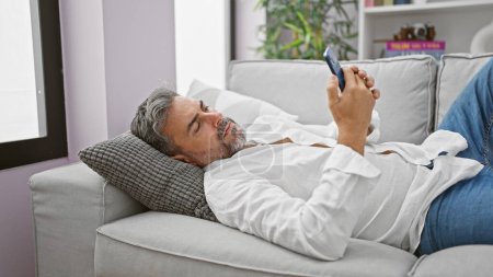 Foto de Joven hombre hispano de pelo gris usando un teléfono inteligente tumbado en un sofá en casa - Imagen libre de derechos