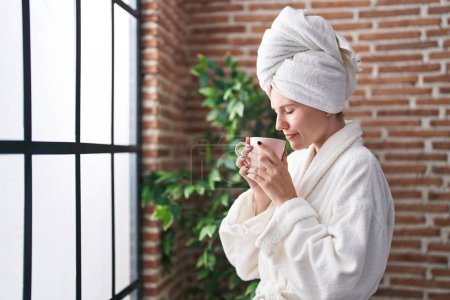 Téléchargez les photos : Jeune femme blonde portant un peignoir sentant le café dans la chambre - en image libre de droit