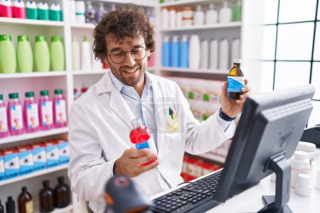 Foto de Joven farmacéutico hispano sonriendo confiado sosteniendo botellas de medicamentos en la farmacia - Imagen libre de derechos