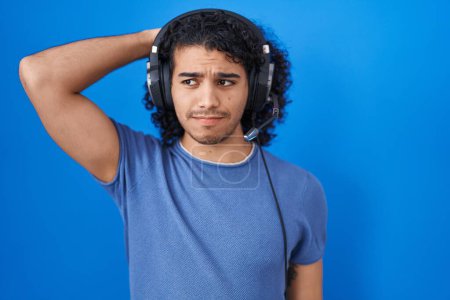 Téléchargez les photos : Homme hispanique aux cheveux bouclés écoutant de la musique à l'aide d'écouteurs confondent et s'interrogent sur la question. incertain avec doute, pensant la main sur la tête. concept coûteux. - en image libre de droit