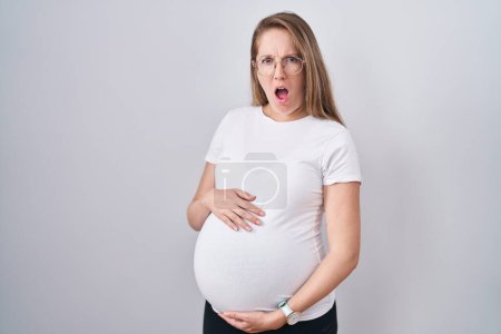 Photo for Young pregnant woman expecting a baby, touching pregnant belly in shock face, looking skeptical and sarcastic, surprised with open mouth - Royalty Free Image