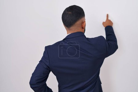 Foto de Joven hombre hispano con traje de negocios y corbata posando hacia atrás apuntando hacia adelante con la mano del dedo - Imagen libre de derechos
