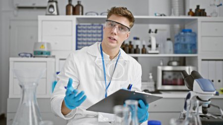 Photo for Attractive, smiling young caucasian male scientist, holding clipboard and passionately speaking about his groundbreaking research in the bustling lab - Royalty Free Image