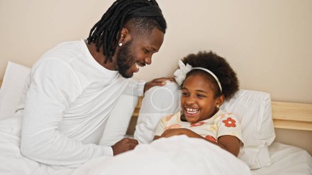 Téléchargez les photos : Afro-américain père et fille couché sur le lit riant beaucoup pour chatouilles dans la chambre - en image libre de droit