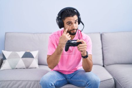 Téléchargez les photos : Jeune homme hispanique jouant contrôleur de tenue de jeu vidéo assis sur le canapé pointant vers l'œil vous regarder geste, expression suspecte - en image libre de droit