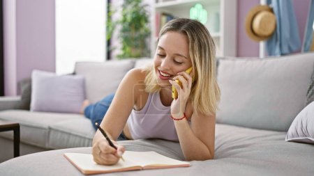 Photo for Young blonde woman writing on notebook speaking on smartphone at home - Royalty Free Image