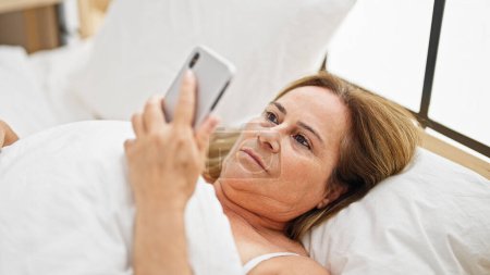 Téléchargez les photos : Moyen Age femme hispanique regardant smartphone couché sur le lit dans la chambre - en image libre de droit