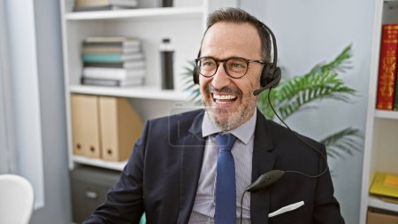 Foto de Sonriente hombre de mediana edad con el pelo gris trabajando su magia en la oficina, con confianza usando un auricular mientras maneja el negocio - Imagen libre de derechos