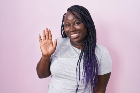 Téléchargez les photos : Jeune femme africaine debout sur fond rose renonçant à dire bonjour heureux et souriant, geste de bienvenue amical - en image libre de droit