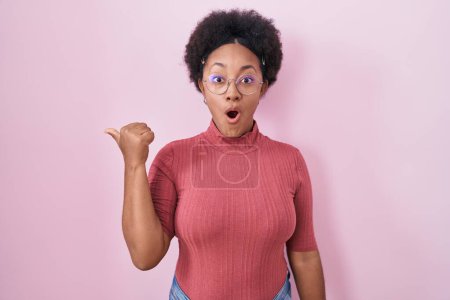 Photo for Beautiful african woman with curly hair standing over pink background surprised pointing with hand finger to the side, open mouth amazed expression. - Royalty Free Image