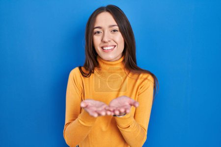 Téléchargez les photos : Jeune femme brune debout sur fond bleu souriant avec les mains paumes ensemble recevant ou donnant geste. maintien et protection - en image libre de droit