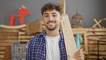 Foto de Sonriendo joven árabe en el trabajo, carpintero profesional maneja con confianza tablón de madera en su estudio de carpintería - Imagen libre de derechos