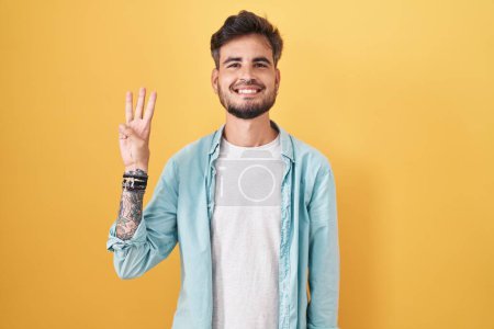 Téléchargez les photos : Jeune homme hispanique avec des tatouages debout sur fond jaune montrant et pointant vers le haut avec les doigts numéro trois tout en souriant confiant et heureux. - en image libre de droit