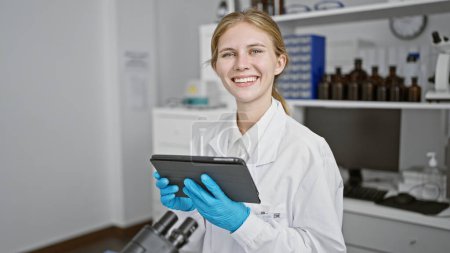 Photo for A young, caucasian, blonde woman wearing a lab coat and gloves smiles while holding a tablet in a laboratory setting. - Royalty Free Image