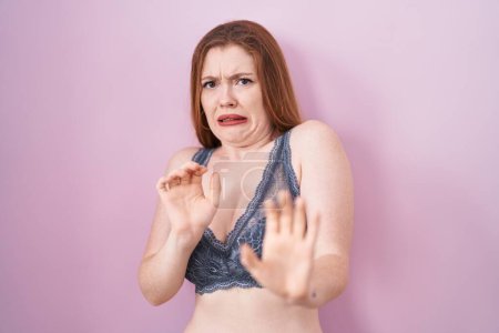 Photo for Redhead woman wearing lingerie over pink background disgusted expression, displeased and fearful doing disgust face because aversion reaction. - Royalty Free Image