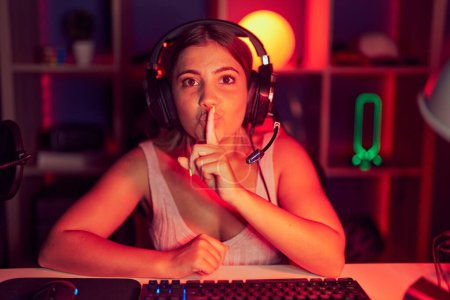 Photo for Young blonde woman playing video games wearing headphones asking to be quiet with finger on lips. silence and secret concept. - Royalty Free Image