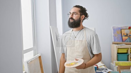 Photo for A thoughtful hispanic man with a beard holds a paint palette in an artist's studio. - Royalty Free Image