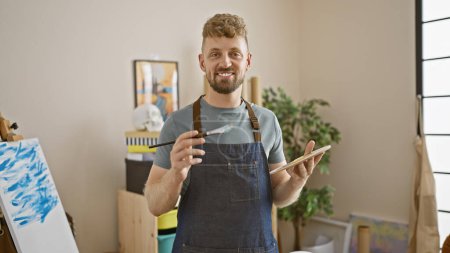 Handsome young caucasian man with blue eyes and a beard smiling in an art studio interior