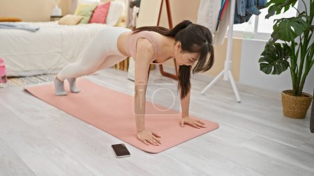 Téléchargez les photos : Femme asiatique pratiquant le yoga dans une chambre lumineuse, sur un tapis rose à côté de son smartphone, exprimant tranquillité et bien-être. - en image libre de droit